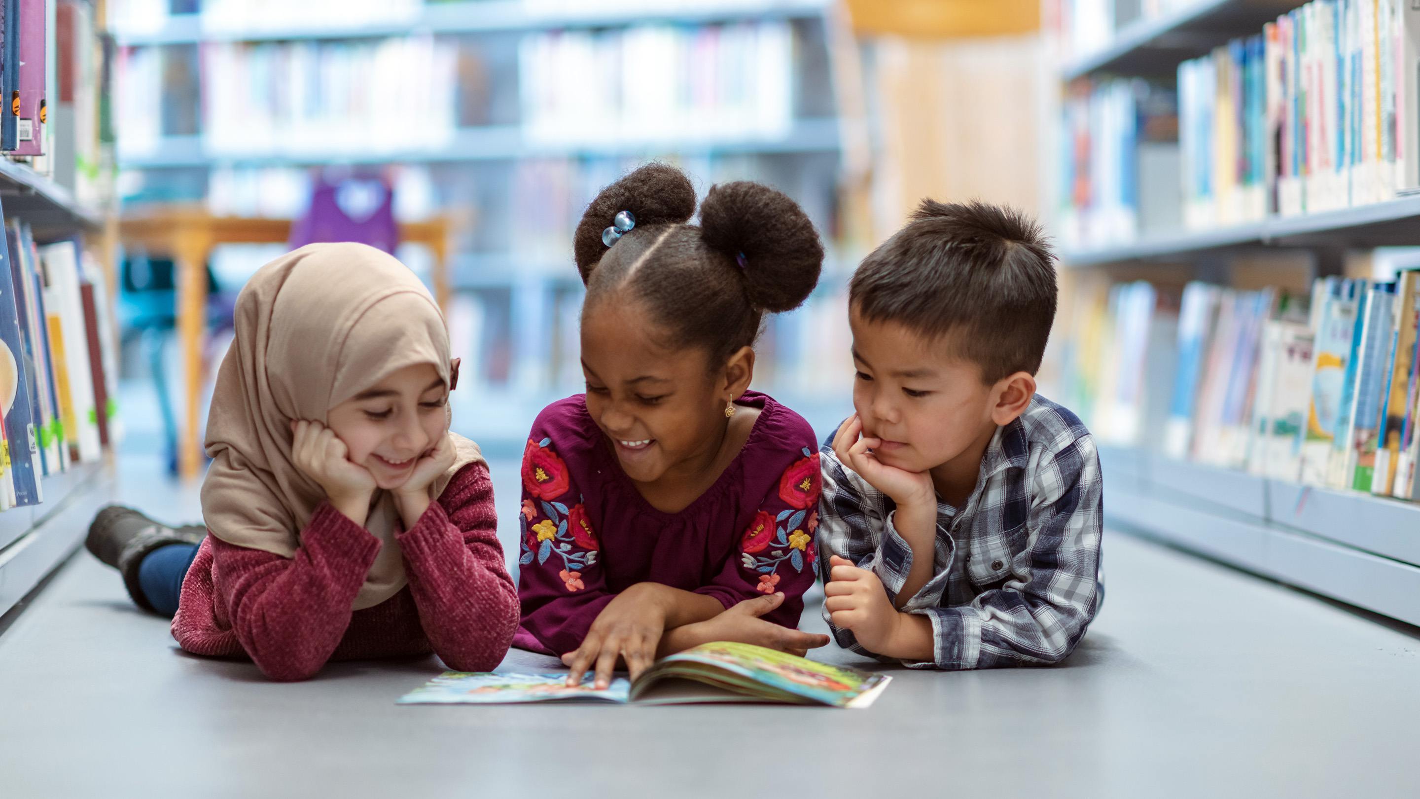 Three small children from different cultures smiling and sitting on the floor as they read a picture book together.