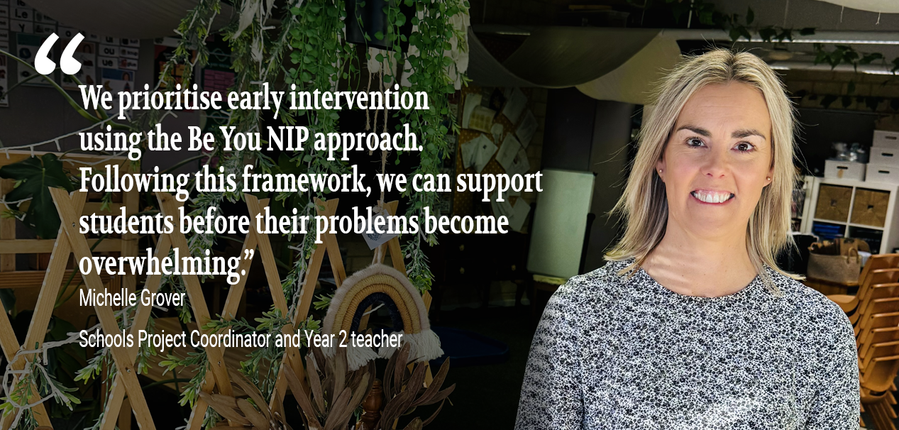 A woman named Michelle Grover, identified as the Schools Project Coordinator and Year 2 teacher, stands smiling in a classroom decorated with plants and natural elements. A quote attributed to her is displayed on the left side of the image: "We prioritise early intervention using the Be You NIP approach. Following this framework, we can support students before their problems become overwhelming.
