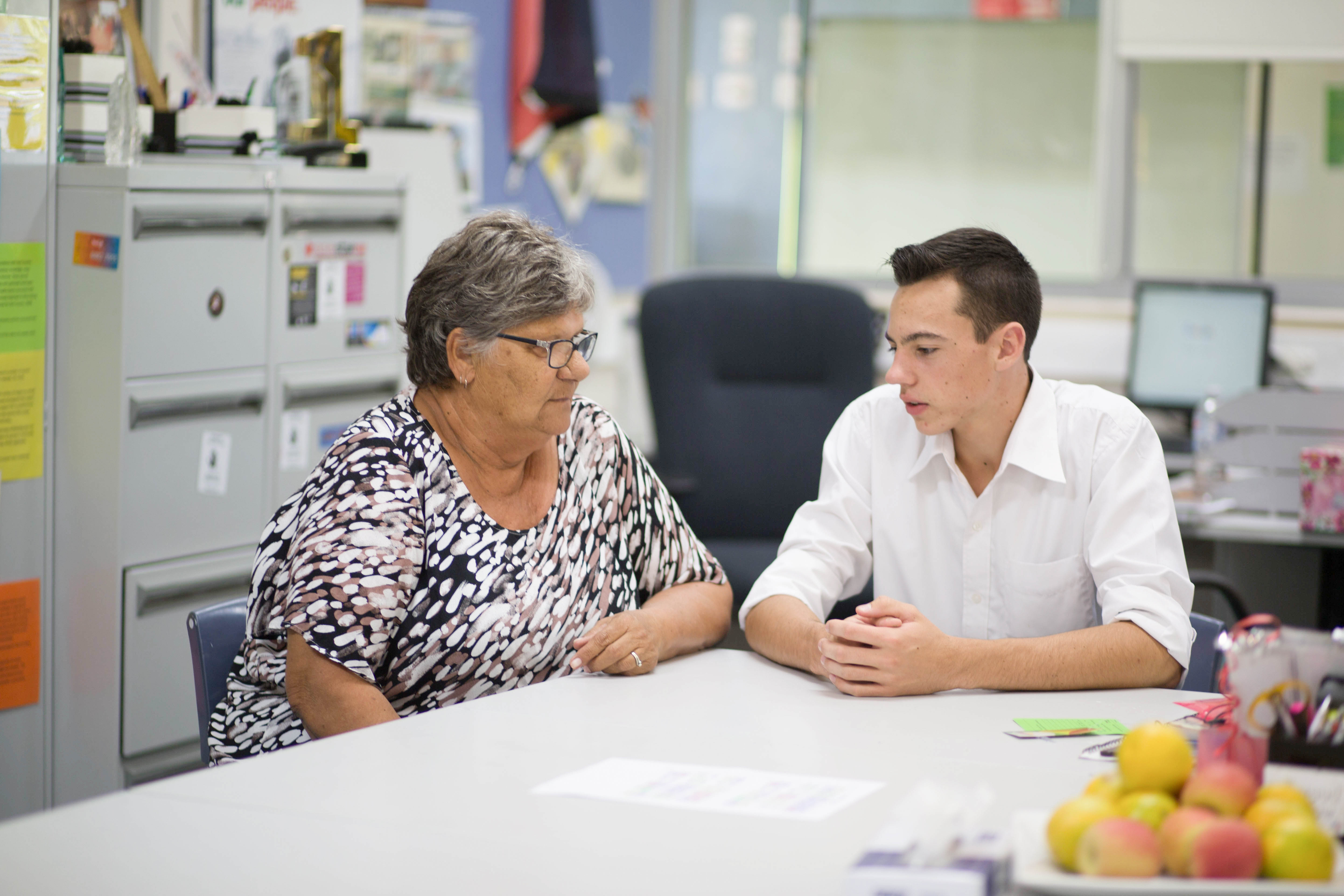 An educator and a secondary aged student are sitting at the corner of a table in an office having a conversation.