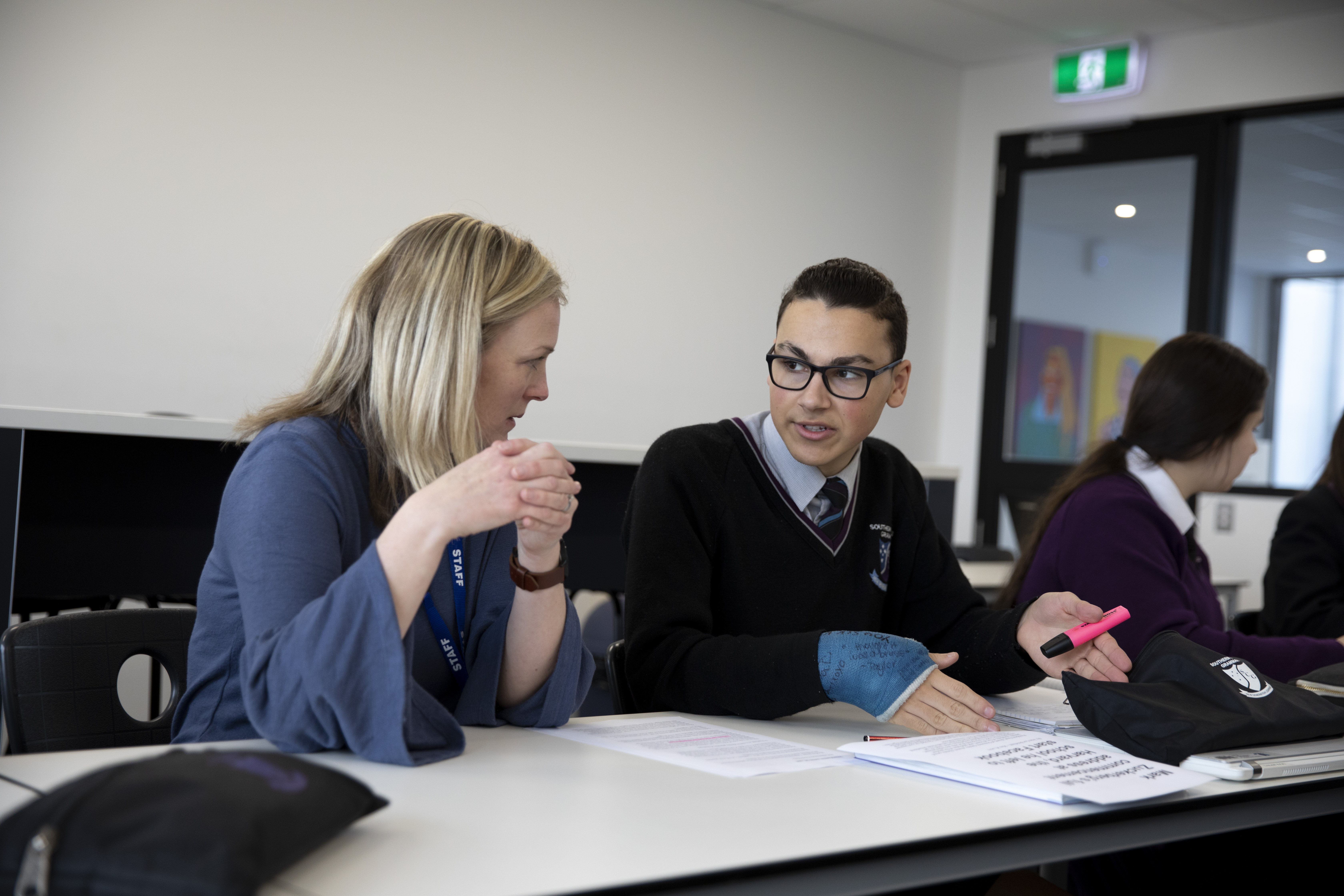 A secondary school student is sitting at a desk talking to the teacher about their work.